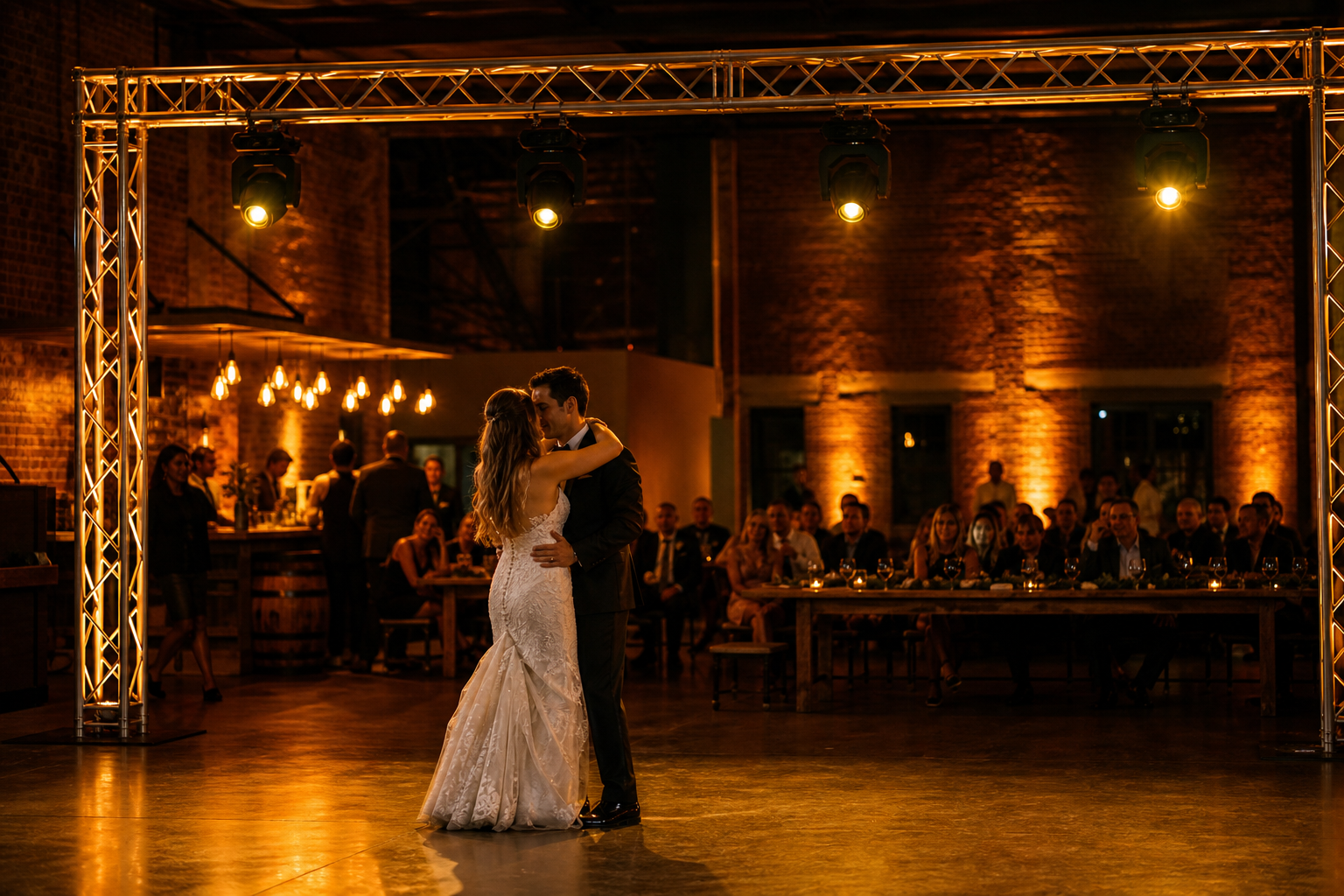 Oakland warehouse wedding reception with exposed brick walls, warm amber uplighting, ground-supported truss with moving heads above the dance floor