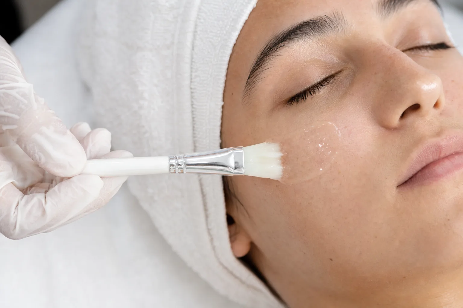 Gloved hand brushing a clear peel solution onto a person's cheek with a soft fan brush, in a clean treatment setting