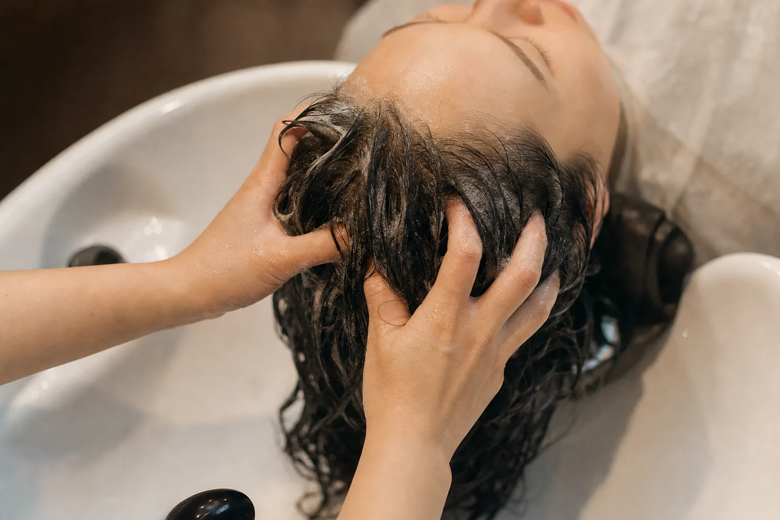 A person reclining at a shampoo basin while a therapist's hands massage their wet scalp, soft warm light
