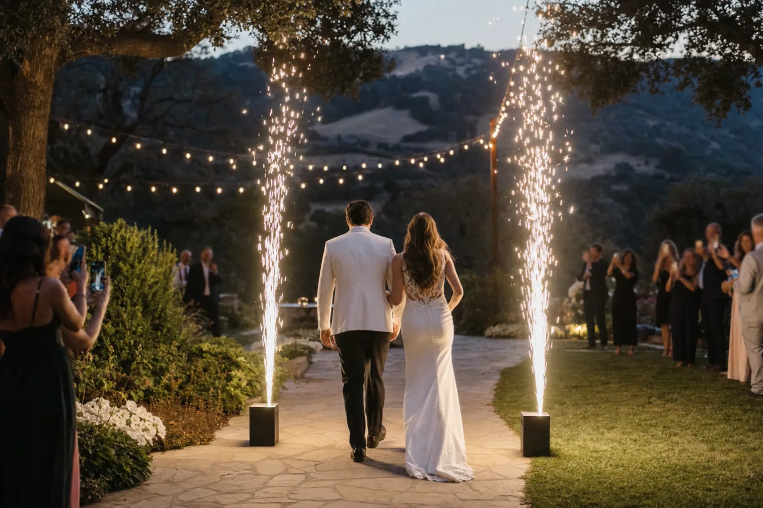 Two cold spark fountains flanking a couple walking out of a garden reception at dusk, with guests watching from a distance