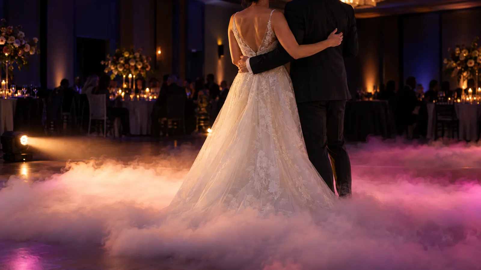 A couple rehearses a first dance as low stage lighting reveals floor-hugging fog against a dark reception room.